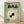 May include: A framed vintage-style poster with sage green background featuring a black goldfish swimming in concentric cream and green circles. Japanese text appears at the top in bold black characters, with smaller text reading "今から" (ima kara). A small illustrated face appears in the bottom left corner. The wooden-framed artwork leans against a white wall on light wooden flooring, with soft natural lighting creating gentle shadows.