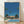 May include: Framed art print featuring a mid-century modern poolside scene with tall palm trees, a colorful building with "PROUD" signage, bright blue swimming pool, and clear blue sky. The artwork displays a retro illustration style with vibrant colors and clean geometric shapes. The print is shown in a light wood frame, leaning against a white textured wall on light wooden flooring with soft natural lighting creating shadows.