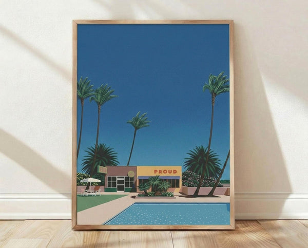 May include: Framed art print featuring a mid-century modern poolside scene with tall palm trees, a colorful building with "PROUD" signage, bright blue swimming pool, and clear blue sky. The artwork displays a retro illustration style with vibrant colors and clean geometric shapes. The print is shown in a light wood frame, leaning against a white textured wall on light wooden flooring with soft natural lighting creating shadows.