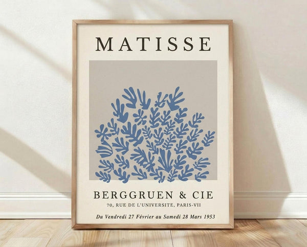 May include: Framed art print featuring blue botanical cutout shapes on beige background, displayed in light wood frame against white textured wall on wooden floor with natural lighting and shadows