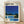 May include: Framed poster print leaning against white wall on light wooden floor. Shows modernist poolside scene with white and yellow mid-century house, bright blue swimming pool, palm trees, and deep blue sky. Yellow lounge chairs beside pool. Cream matting in natural wood frame. Japanese text and "Pacific Breeze Collection Tokyo, 1982" at top with colorful logo.