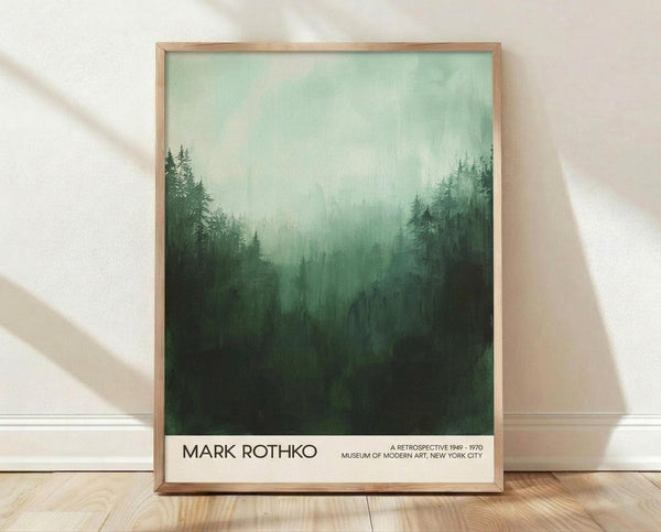 May include: Framed poster displaying misty forest landscape in green tones that fade from dark evergreen trees at bottom to pale mint sky at top. Text reads "MARK ROTHKO A RETROSPECTIVE 1949-1970 MUSEUM OF MODERN ART, NEW YORK CITY" in black letters on white bottom margin. Natural wood frame leans against white textured wall on light wooden floor with soft shadows.