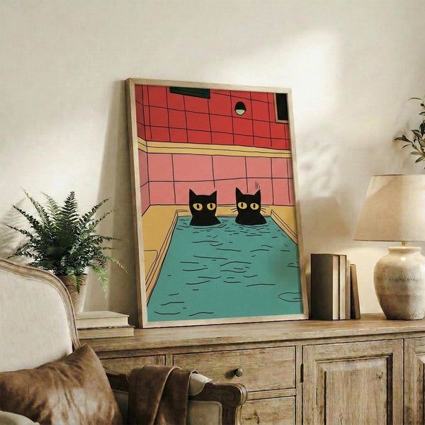 May include: Framed poster print showing two black cats with yellow eyes sitting in a turquoise swimming pool, set against red-pink tiled bathroom walls. The artwork has a mid-century modern illustration style with bold colors and geometric patterns. The print is displayed on a rustic wooden sideboard alongside books, a ceramic vase, and a potted fern plant in a cozy living room setting with warm lighting.