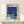 May include: Framed vintage-style poster featuring a modernist poolside scene with glass house, bright blue swimming pool, and palm trees against deep blue sky, displayed on light wood shelf beside small wrapped gift with dried grass decoration