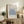 May include: Framed Matisse exhibition poster featuring blue coral-like botanical cutout design on beige background, displayed on rustic wooden sideboard alongside fern plant, ceramic vases, and books in warm neutral living room setting with beige armchair visible