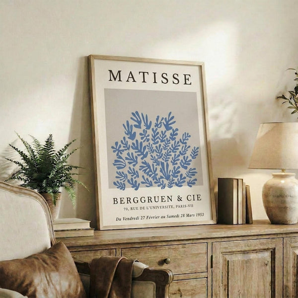 May include: Framed Matisse exhibition poster featuring blue coral-like botanical cutout design on beige background, displayed on rustic wooden sideboard alongside fern plant, ceramic vases, and books in warm neutral living room setting with beige armchair visible