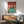 May include: Framed poster print displaying two black cats with yellow eyes sitting in a turquoise swimming pool inside a pink-tiled bathroom. The artwork features cartoon-style illustration with bold colors. Wood-framed credenza below holds a white ceramic table lamp. Mid-century modern mint green upholstered chair with wooden arms sits nearby. Warm sunlight casts shadows across white walls in this styled room mockup scene.