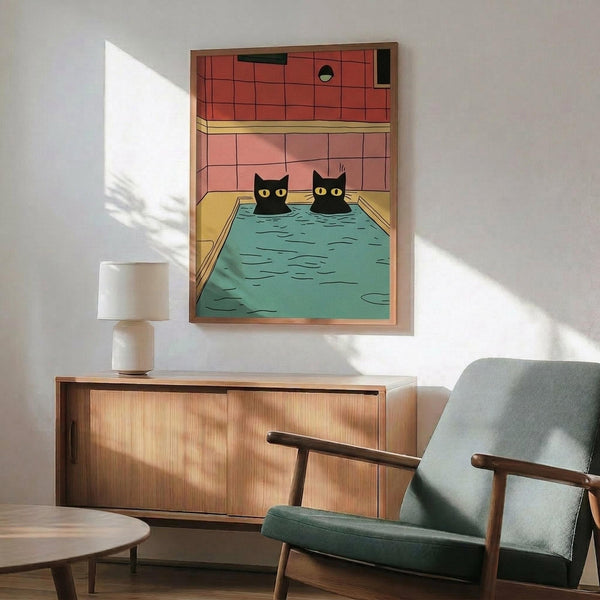 May include: Framed poster print displaying two black cats with yellow eyes sitting in a turquoise swimming pool inside a pink-tiled bathroom. The artwork features cartoon-style illustration with bold colors. Wood-framed credenza below holds a white ceramic table lamp. Mid-century modern mint green upholstered chair with wooden arms sits nearby. Warm sunlight casts shadows across white walls in this styled room mockup scene.