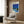 May include: Framed poster showing modernist house with swimming pool under blue sky, displayed on wood paneled wall above beige upholstered armchair with geometric patterned pillow. Gold floor lamp and dark round side table with open book visible. Poster features clean lines, bright blues, and mid-century architectural style in minimalist composition.