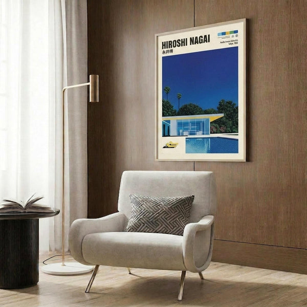 May include: Framed poster showing modernist house with swimming pool under blue sky, displayed on wood paneled wall above beige upholstered armchair with geometric patterned pillow. Gold floor lamp and dark round side table with open book visible. Poster features clean lines, bright blues, and mid-century architectural style in minimalist composition.