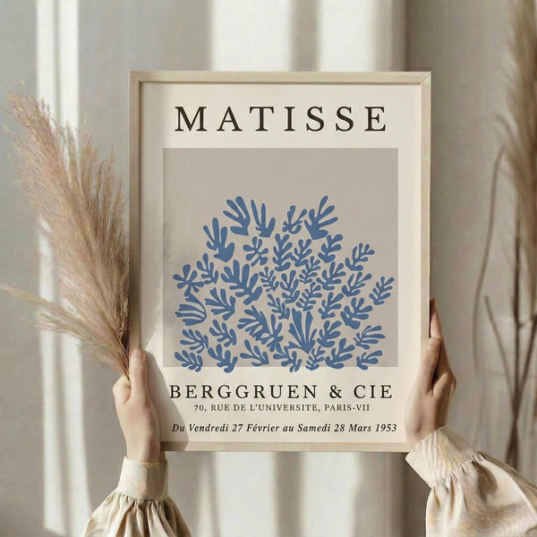 May include: Hands holding a framed art print featuring blue coral-like botanical shapes on beige background with "MATISSE" and "BERGGRUEN & CIE" text. Light wood frame, neutral curtain backdrop, dried pampas grass visible to left side.