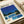 May include: Framed art print displayed on white bedding showing a mid-century modern poolside scene with geometric architectural buildings, bright blue swimming pool, lush green palm trees and foliage against a deep blue sky. The minimalist style features clean lines and bold colors in yellow, blue, and white. A white picture frame surrounds the artwork, positioned next to rolled white paper or fabric on soft bedding.