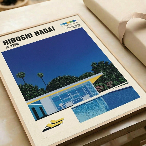 May include: Framed art print displayed on white bedding showing a mid-century modern poolside scene with geometric architectural buildings, bright blue swimming pool, lush green palm trees and foliage against a deep blue sky. The minimalist style features clean lines and bold colors in yellow, blue, and white. A white picture frame surrounds the artwork, positioned next to rolled white paper or fabric on soft bedding.