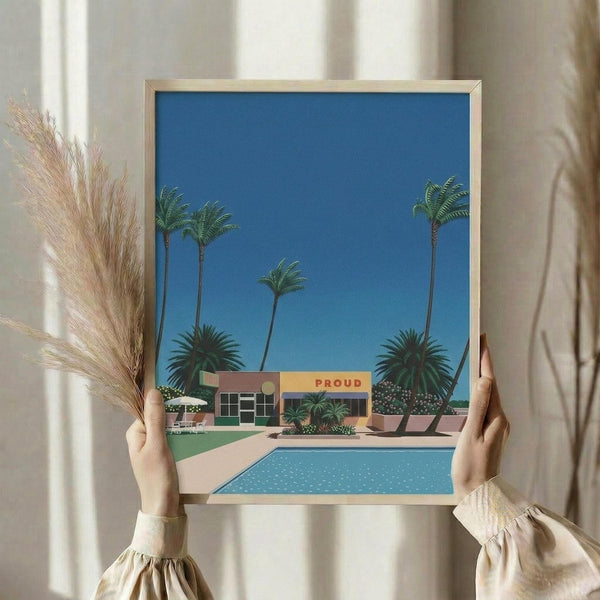 May include: Hands holding a framed artwork showing a mid-century modern house with swimming pool, tall palm trees against bright blue sky, and "PROUD" text on the building. The print has a vintage illustration style with clean geometric shapes and vibrant colors. Person wears cream-colored clothing with gathered sleeves, and dried pampas grass appears in the background near white curtains.