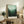 May include: Framed forest landscape poster with misty green trees fading from dark forest floor to pale mint sky, displayed on rustic wooden sideboard beside fern plant, cream ceramic vase, and stacked books in neutral-toned living room with beige upholstered furniture.