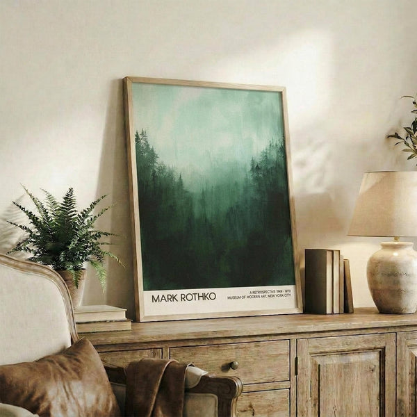 May include: Framed forest landscape poster with misty green trees fading from dark forest floor to pale mint sky, displayed on rustic wooden sideboard beside fern plant, cream ceramic vase, and stacked books in neutral-toned living room with beige upholstered furniture.