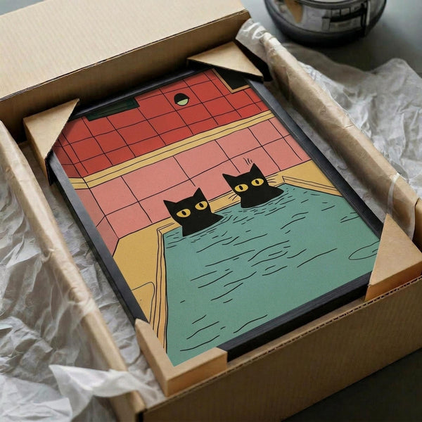 Framed poster artwork featuring two black cats with yellow eyes peeking out from a turquoise bathtub in a retro-style bathroom with coral pink tiled walls. The illustration has a vintage comic book aesthetic with bold colors and clean lines. The black-framed print is shown in an opened cardboard shipping box with protective tissue paper wrapping on a white surface.