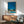 May include: Mid-century modern living room featuring a framed poster of a modernist poolside scene with palm trees against a bright blue sky. The artwork shows a pink building with "PROUD" signage and turquoise swimming pool. Displayed above a wooden sideboard with white ceramic lamp, next to a sage green upholstered armchair with wooden frame. Natural sunlight creates geometric shadows on white wall.