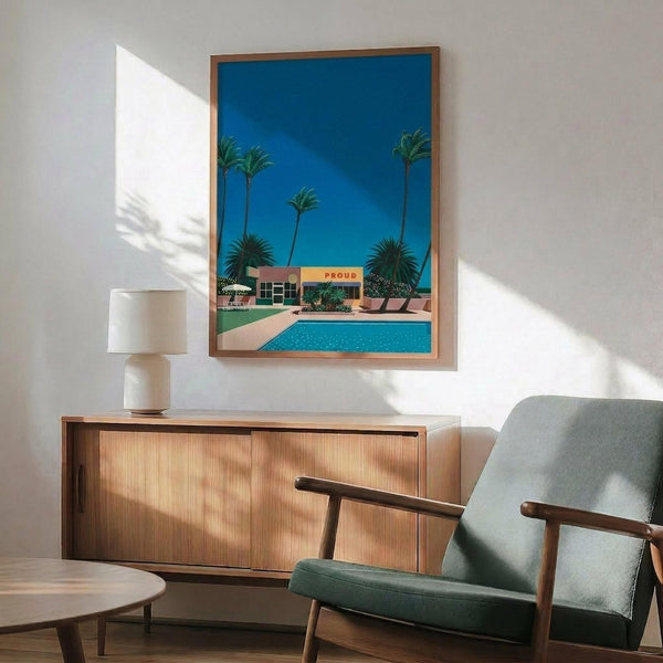 May include: Mid-century modern living room featuring a framed poster of a modernist poolside scene with palm trees against a bright blue sky. The artwork shows a pink building with "PROUD" signage and turquoise swimming pool. Displayed above a wooden sideboard with white ceramic lamp, next to a sage green upholstered armchair with wooden frame. Natural sunlight creates geometric shadows on white wall.