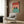 May include: Framed poster showing two black cats with yellow eyes sitting in a turquoise bathtub surrounded by coral pink subway tile walls. Modern minimalist room setting with beige upholstered armchair, geometric throw pillow, brass floor lamp, dark round side table with open book, and wood panel wall. Clean contemporary interior design style.