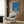May include: Retro-style poster print featuring a mid-century modern poolside scene with palm trees, blue sky, and turquoise swimming pool, displayed in light wood frame on brown wood-paneled wall above cream upholstered armchair with geometric patterned pillow, next to brass floor lamp and round side table with open book.