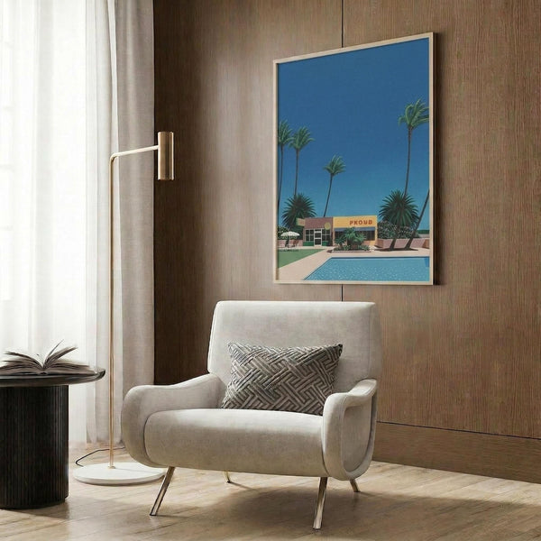 May include: Retro-style poster print featuring a mid-century modern poolside scene with palm trees, blue sky, and turquoise swimming pool, displayed in light wood frame on brown wood-paneled wall above cream upholstered armchair with geometric patterned pillow, next to brass floor lamp and round side table with open book.
