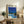 May include: Framed poster featuring a modernist house with swimming pool under bright blue sky with palm trees, displayed on rustic wooden sideboard alongside potted fern, ceramic vase, and stacked books in neutral-toned living room with beige upholstered furniture.