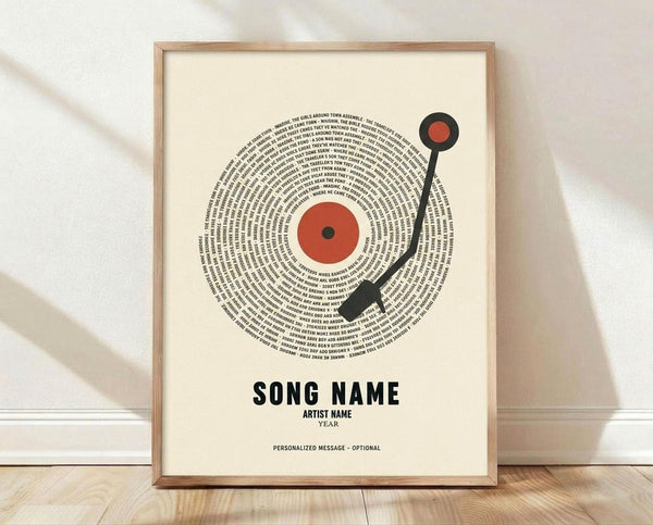 May include: Framed poster print featuring a vinyl record design with song lyrics arranged in concentric circles around a red center label and black tonearm. The cream-colored artwork displays placeholder text reading "SONG NAME," "ARTIST NAME," "YEAR," and "PERSONALIZED MESSAGE - OPTIONAL" in black typography. Light wood frame sits on wooden flooring against white textured wall with soft natural lighting and shadows.