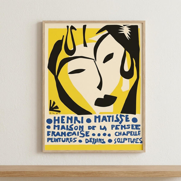 May include: Framed vintage exhibition poster with yellow background featuring stylized black and cream faces in Matisse's cut-out style, blue French text below, displayed on white wall above light wood surface in minimalist room setting.