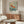 May include: Framed poster featuring a golden-orange koi fish swimming among cherry blossoms in Japanese art style, displayed above a mid-century modern walnut credenza in a minimalist living room. White upholstered chair and glass coffee table visible in foreground, with modern brass floor lamp creating warm lighting on neutral walls.