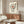 May include: Modern living room with beige walls featuring framed vinyl record poster art with red center label, black tonearm, and spiral text design. Wood credenza below holds white sculptural lamp. White curved chair and glass coffee table in foreground. Minimalist Scandinavian-style interior with warm wood tones and neutral palette.