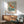 May include: A framed vintage Japanese art print featuring a golden-orange koi fish swimming among cherry blossoms in muted blue-green water. The poster hangs above a mid-century modern wood credenza with sliding doors, next to a ceramic table lamp. A teal upholstered armchair with wooden frame sits nearby on a round area rug. Natural sunlight creates geometric shadows on the white wall.