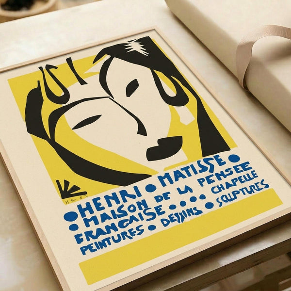 A framed vintage poster print featuring a stylized portrait of a woman with black hair against a bright yellow background, rendered in Matisse's distinctive cut-out style. Blue French text appears at the bottom. The cream-colored frame sits on a light wooden surface next to rolled white papers, creating soft shadows in natural lighting.