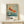 May include: Framed vintage Japanese art poster featuring orange and red koi fish swimming among cherry blossoms in blue-green water. Exhibition poster shows museum text and dates below artwork. Natural wood frame sits on light wood surface next to small wrapped gift with dried branch. Soft shadows cast on cream wall behind.