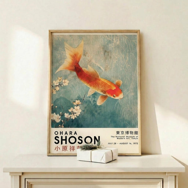 May include: Framed vintage Japanese art poster featuring orange and red koi fish swimming among cherry blossoms in blue-green water. Exhibition poster shows museum text and dates below artwork. Natural wood frame sits on light wood surface next to small wrapped gift with dried branch. Soft shadows cast on cream wall behind.