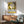 May include: Framed Henri Matisse exhibition poster with yellow background featuring stylized black and white faces, displayed above a mid-century modern wooden credenza with table lamp. Sage green upholstered chair with wooden arms sits nearby on hardwood floor. Warm sunlight creates dramatic shadows on white wall in contemporary living room setting.