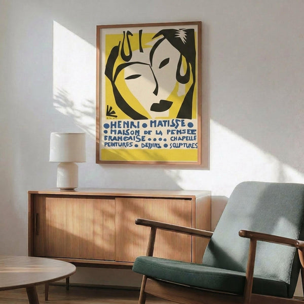 May include: Framed Henri Matisse exhibition poster with yellow background featuring stylized black and white faces, displayed above a mid-century modern wooden credenza with table lamp. Sage green upholstered chair with wooden arms sits nearby on hardwood floor. Warm sunlight creates dramatic shadows on white wall in contemporary living room setting.