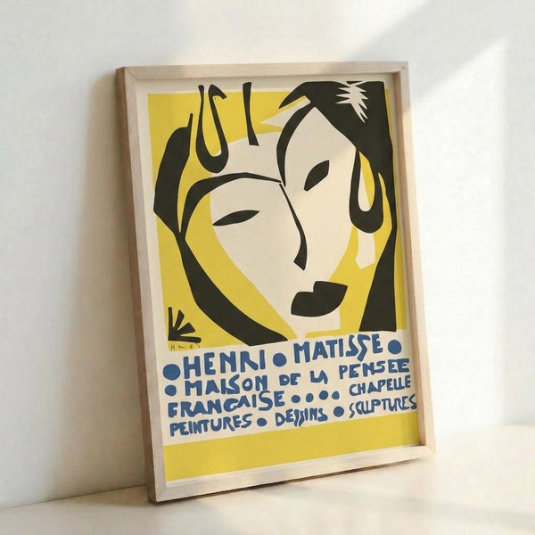 Framed vintage exhibition poster featuring a stylized face illustration in black, cream and yellow. The minimalist design shows a woman's profile with closed eyes and flowing hair in Matisse's signature cut-out style. Blue text reads "HENRI MATISSE MAISON DE LA PENSEE FRANCAISE CHAPELLE PEINTURES DESSINS SCULPTURES" against a bright yellow background. The poster is displayed in a light wood frame leaning against a white wall.