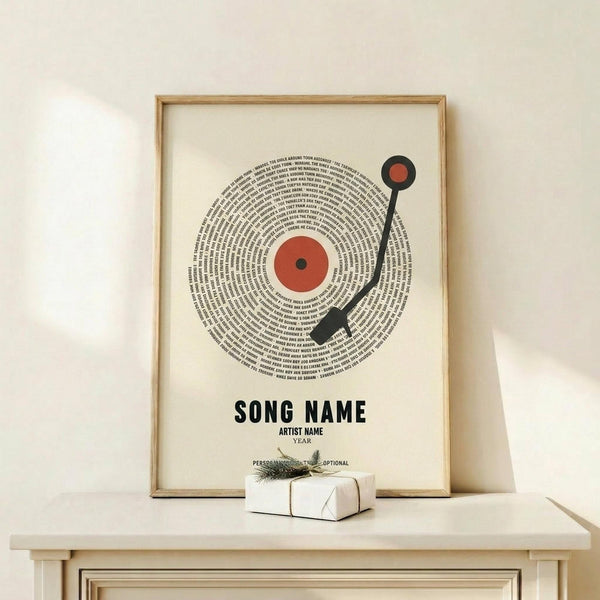 May include: Framed poster displaying a vinyl record design with turntable arm, featuring song lyrics arranged in concentric circles around an orange center label. Black text reads "SONG NAME," "ARTIST NAME," and "YEAR" below the record illustration. Light wood frame sits on natural wood shelf beside small white gift box with evergreen sprig. Cream colored wall background with soft natural lighting.