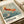 May include: Vintage exhibition poster featuring a golden-orange koi fish swimming among delicate white cherry blossoms against a muted blue-green background. The artwork shows Japanese woodblock print styling with fine linear details. Text at bottom reads "OHARA SHOSON" with Japanese characters and museum exhibition dates from 1972. Poster displayed on cream bedding with neutral ribbon visible.