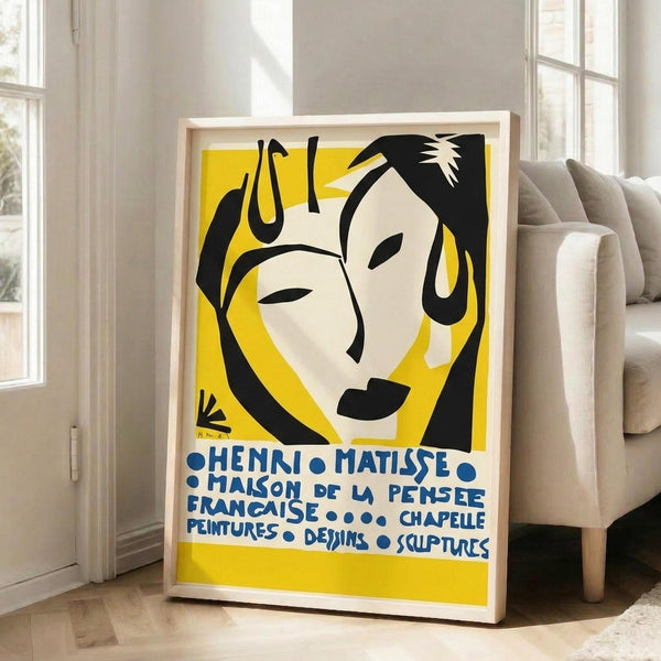 May include: Framed Henri Matisse exhibition poster with yellow background featuring black silhouettes of two faces in his characteristic cut-out style. Blue text in French reads "HENRI MATISSE MAISON DE LA PENSEE FRANCAISE CHAPELLE PEINTURES DESSINS SCULPTURES." Light wood frame leans against white wall next to beige upholstered sofa in bright, minimalist living room with large windows.