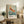 May include: Framed exhibition poster featuring a golden-red koi fish swimming among delicate cherry blossoms on a soft blue-green background, displayed on a rustic wooden console table alongside fern plants, ceramic vases, and books in a warm, neutral-toned living room setting.