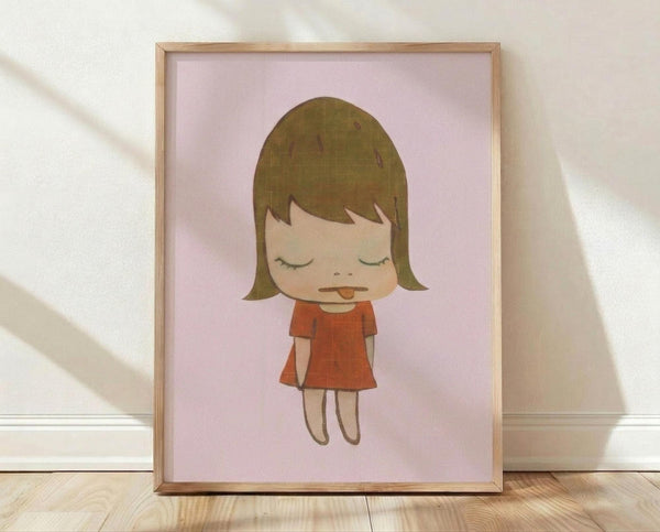 May include: Framed art print featuring a stylized cartoon girl with olive green bob haircut and closed eyes, wearing an orange-red dress, set against a soft pink background. The minimalist illustration shows the character with a small tongue sticking out. Light wooden frame sits on wooden flooring against white walls with natural window light creating gentle shadows.