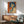 May include: Framed vintage-style poster featuring woman with black bob haircut holding gun against orange background with Japanese text, displayed above wooden sideboard with white ceramic lamp, next to sage green mid-century armchair in bright sunlit room with white walls
