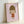 May include: Framed art print featuring a cartoon-style illustration of a young girl with brown bob haircut and closed eyes, wearing an orange-red dress, sticking out her tongue playfully. The minimalist artwork is displayed against a soft pink background within a light wood frame, positioned on what appears to be a white surface against a white wall.