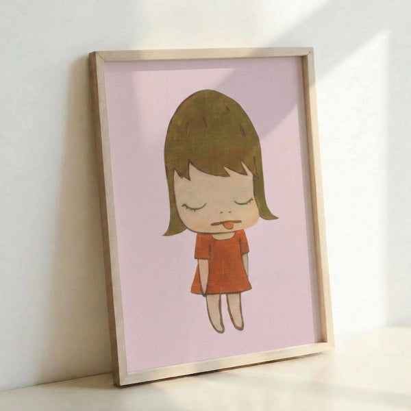 May include: Framed art print featuring a cartoon-style illustration of a young girl with brown bob haircut and closed eyes, wearing an orange-red dress, sticking out her tongue playfully. The minimalist artwork is displayed against a soft pink background within a light wood frame, positioned on what appears to be a white surface against a white wall.