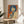 May include: Framed vintage-style Japanese poster featuring a woman with black bob haircut holding a gun, rendered in orange and blue tones with Japanese text, displayed on a round wooden side table next to a beige upholstered chair with decorative pillow and plant leaves visible in soft natural lighting