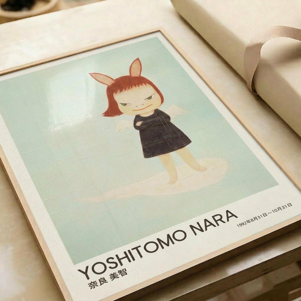 May include: Framed art print featuring a stylized illustration of a child-like figure with reddish-brown hair and bunny ears, wearing a dark dress, rendered in soft pastels against a pale mint green background. The white matted frame sits on light wooden surfaces with rolled paper nearby, showing Japanese text and exhibition details at the bottom of the print.