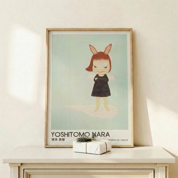 May include: Framed art print featuring a stylized character with reddish-brown bob haircut, bunny ears, and dark dress against mint green background. The poster shows "YOSHITOMO NARA" text at bottom with Japanese characters and dates. Displayed in light wood frame on white wooden shelf beside small wrapped gift with evergreen sprig, against cream colored wall.