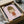 May include: Framed art print featuring a cute cartoon-style girl with brown bob haircut and closed eyes, wearing an orange dress, set against a soft pink background. The print is displayed in a black frame with cardboard packaging materials and wooden frame corners visible around it on a white surface.